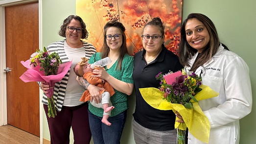 Four women stand against a green wall with an orange flowered photo in the background. The woman second from the left is holding her newborn baby girl. Her partner stands to her left. The two medical providers are standing on the outside of each parent on either side, and are holding flowers from the parents..