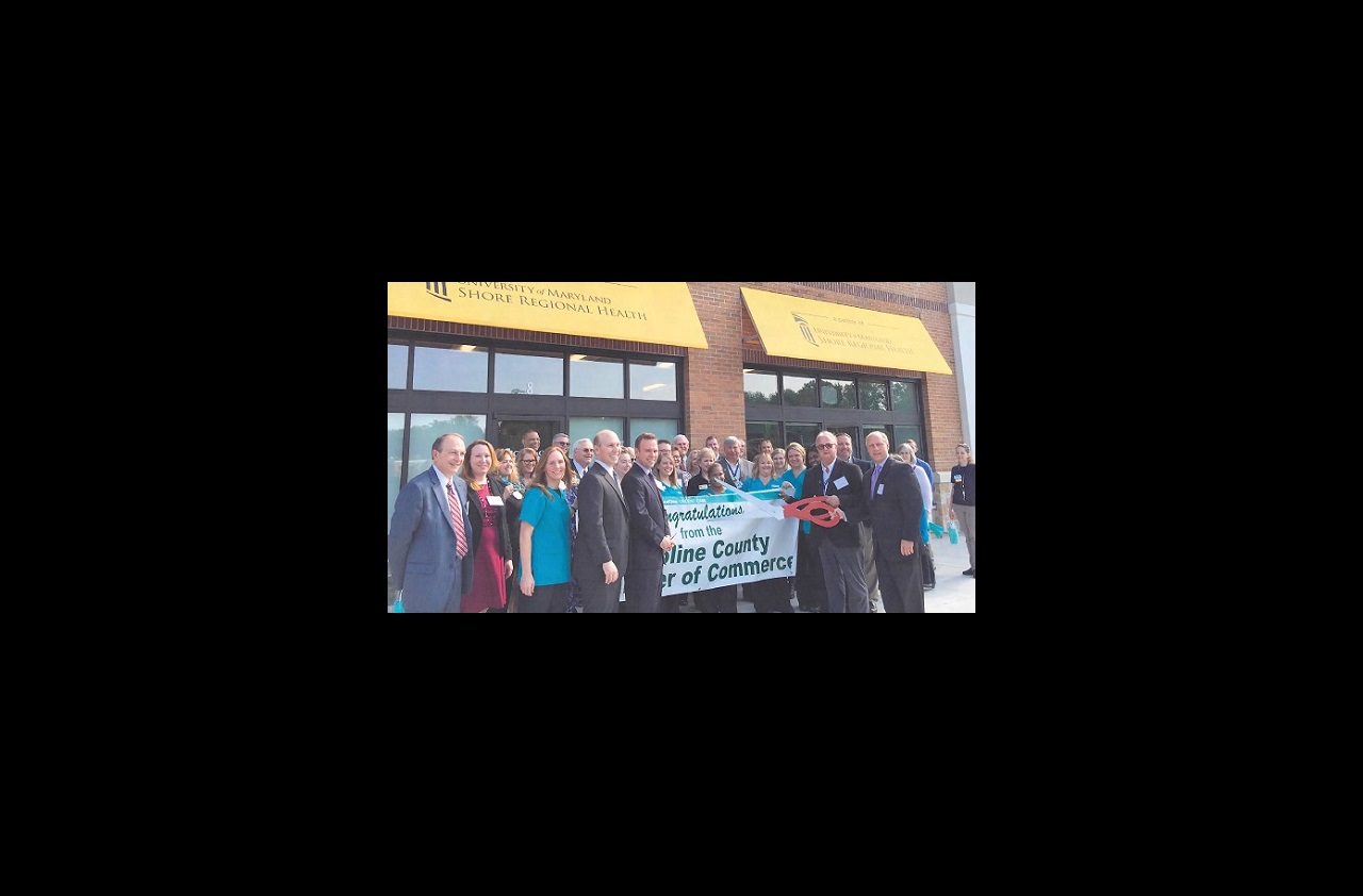 A group of people gather in front of the urgent care location in Denton, Maryland. Ken Kozel and Larry Porter hold a large pair of scissors and many people are helping to hold a banner congratulating the people of Caroline County, from the Caroline County Chamber of Commerce.