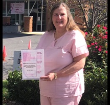 A woman in pink scrubs smiles in front of some trees, holding a sign about Breast Cancer Awareness Month.