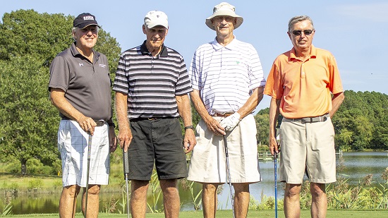 Four men stand in front of a lake, holding golf clubs, on a golf course. It is sunny and they are dressed for summer.
