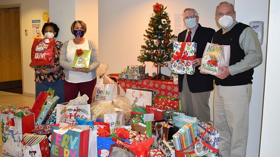 Two women stand to the left of a Christmas tree and two men stand to the right. They are holding Christmas gifts and the floor around the tree is piled high with Christmas gifts.