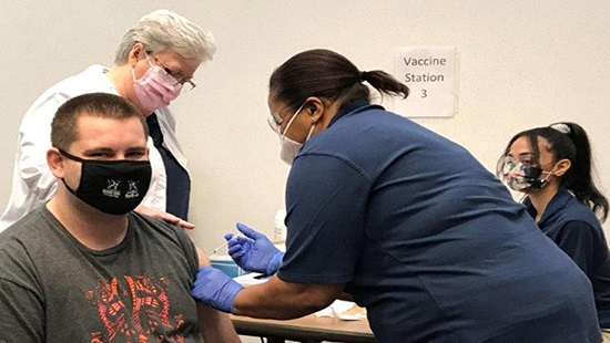 A man is shown getting the COVID-19 vaccine from two female students as a female nursing educator watches.