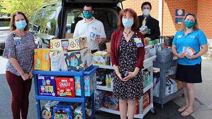 Five people stand in front of the back of a car with a lot of food donations stacked on rolling carts.