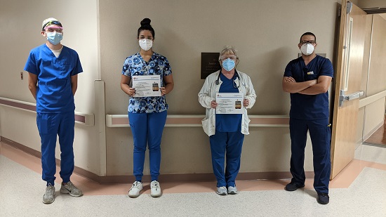 Four people stand socially distanced, smiling, and one woman in the middle is holding a sign indicating they won an award for Stroke Care from the American Heart Association.