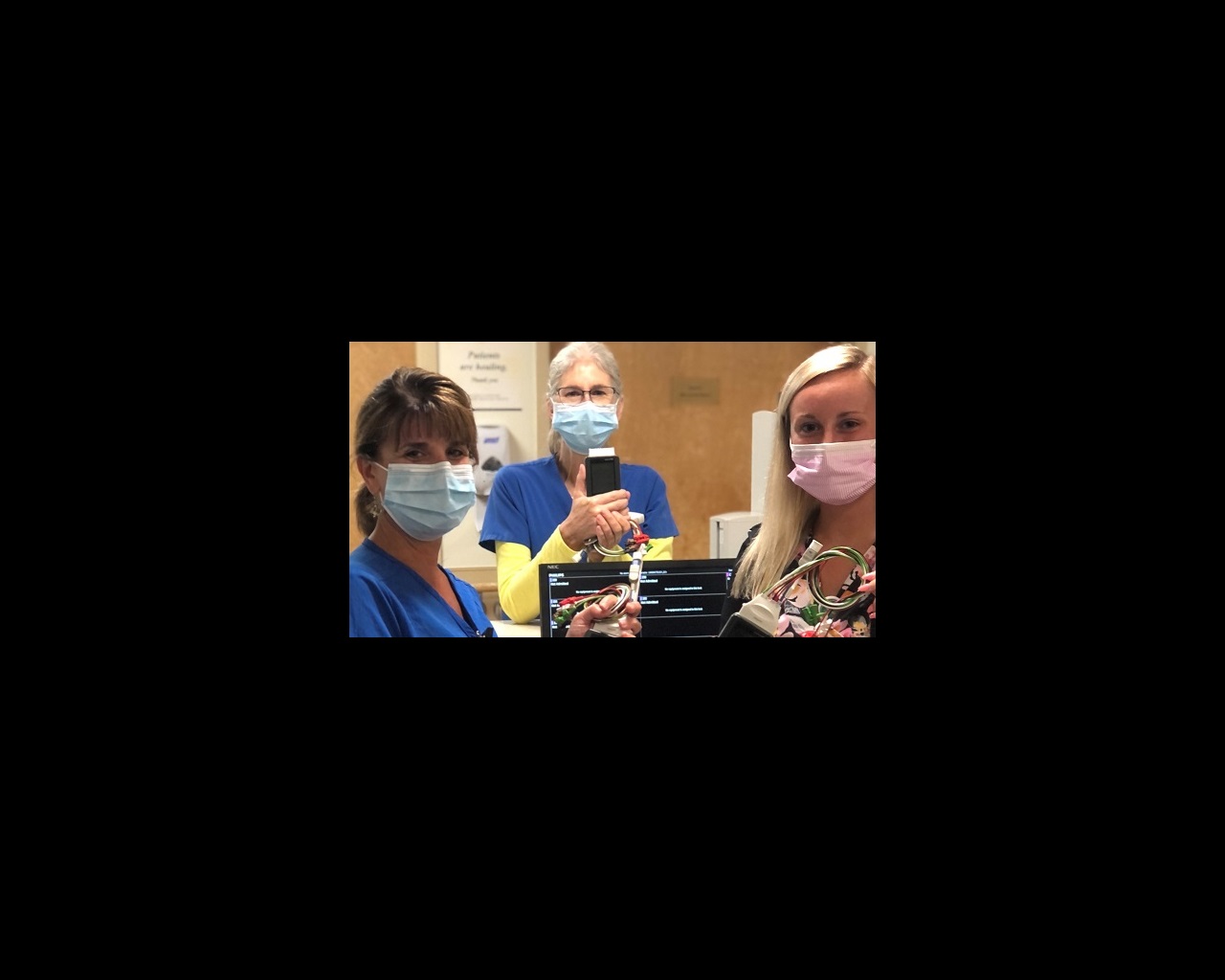 Three women are standing and showing off new handheld hospital critical care equipment. They are smiling behind their masks.