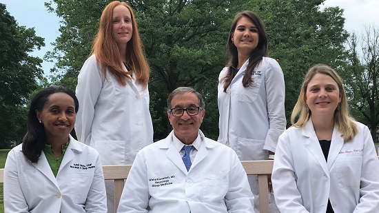 Three neurology team members are sitting on a bench. Two women flanking one man. Two other female team members are standing up behind them, behind the bench.