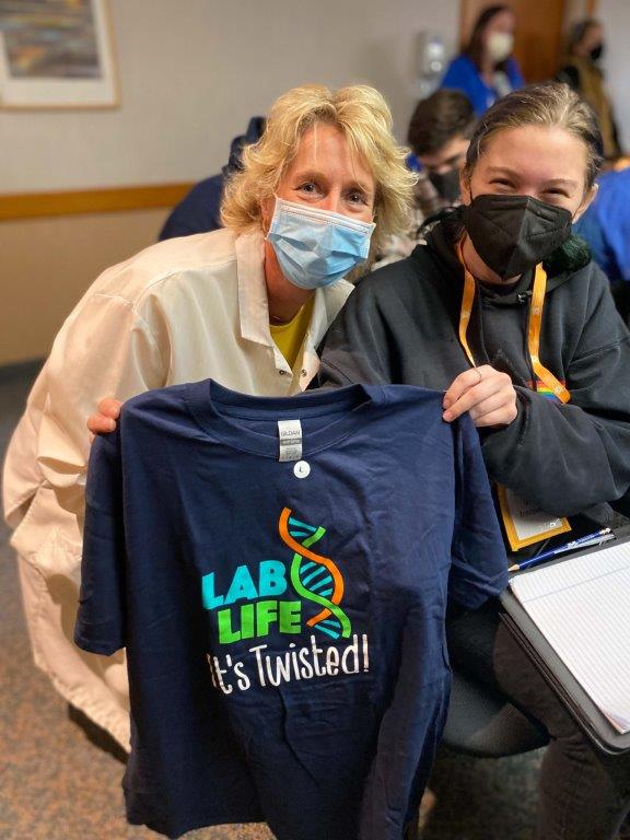 Two women who are masked are posing for a photo, holding a t-shirt that says "Lab Life."