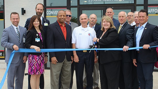 A group of people stand in front of the front doors of the new UM Urgent Care location in Chester, Maryland, on Kent Island. They are holding a blue ribbon and are smiling at the camera before the blue ribbon is cut.