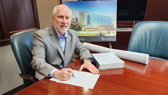 Ken Kozel sits at a desk with a rendering of the new regional medical center in the background. He is signing the Certificate of Need document.