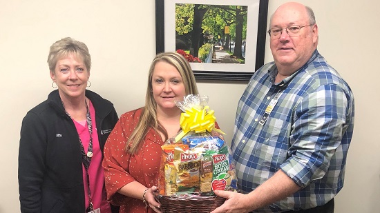 A woman stands in the center of a photo holding a gift basket. She has a UM SRH team member to her left, a woman, and to her right, a man, from the Population Health team.
