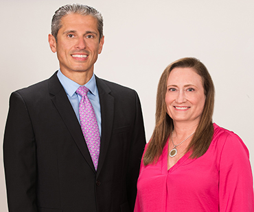 Dr. Kurtom and Wendy Towers stand next to each other smiling. Dr. Kurtom is wearing a dark suit with a pink tie and Wendy is wearing a pink blouse.