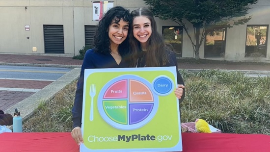 Two women standing behind a table, holding a MyPlate chart.