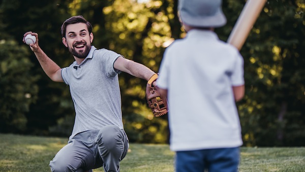 Man squats to throw baseball to child holding a baseball hat