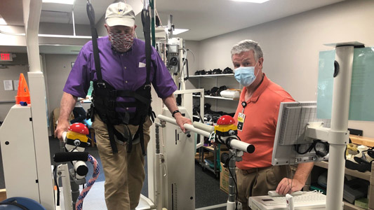 A man in a purple shirt uses Lokomat technology during a rehabilitation appointment, with his instructor in an orange shirt looking at the camera. There is other rehabilitation equipment around the room.